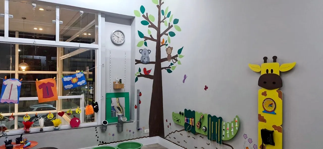 Sala de estimulação psicomotora com uma árvore pintada na parede, painéis de atividades em forma de jacaré e de girafa e um varal de roupinhas de feltro