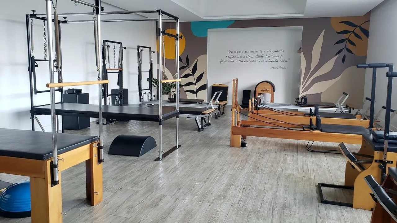 Estúdio de pilates com cadillacs, reformers e chair diante de um mural de folhas com uma frase de Joseph Pilates